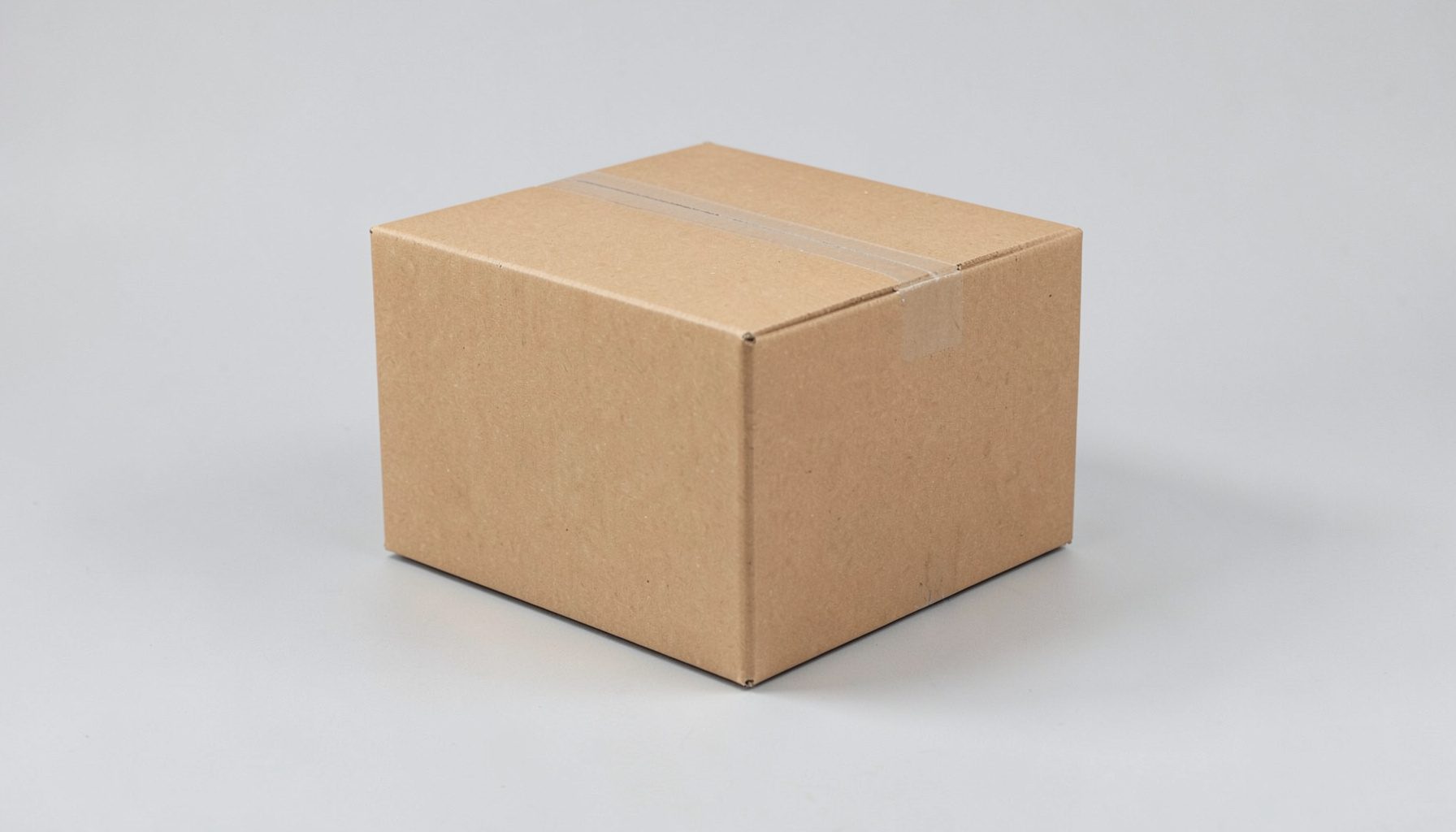 Plain cardboard shipping box on a neutral light gray surface, clean and minimal composition with soft shadows